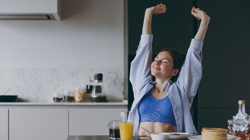 Žena se proteže za stolom u kuhinji dok ispred nje stoji doručak sa sokom, palačinkama i voćem